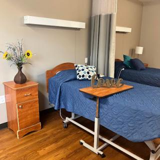 A well-organized hospital or nursing home room featuring two beds with blue bedding, a bedside table with a decorative vase, and a tray for meals or personal items.