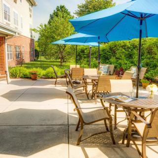 A sunny patio with tables, chairs, and blue umbrellas surrounded by greenery.