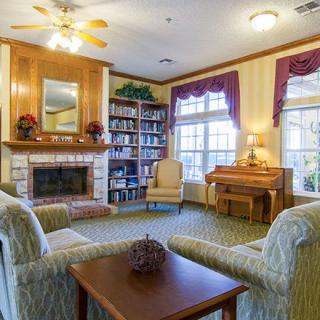 A cozy living room with a fireplace and bookshelves, featuring armchairs and a piano by the window.