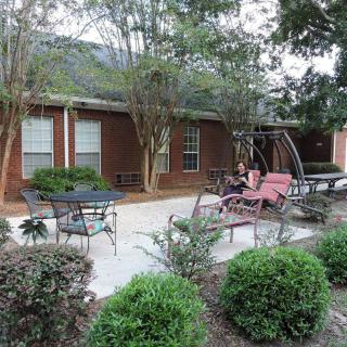 A cozy patio area with outdoor furniture surrounded by trees and bushes next to a brick building.