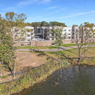 A serene view of modern apartments adjacent to a tranquil lake, surrounded by lush greenery.