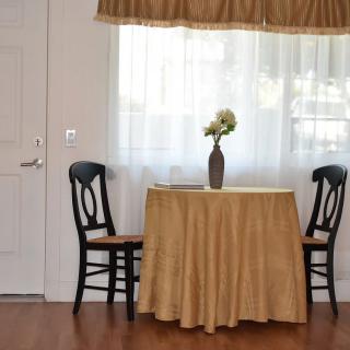 A small round table with a golden tablecloth is set between two black chairs, decorated with a vase of flowers, in front of a window with white curtains.