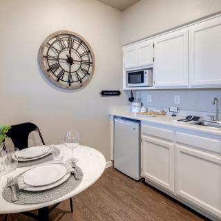 A small minimalist kitchen with white cabinets, a round dining table set for two, and a large decorative clock on the wall.