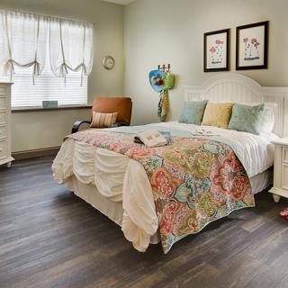 A cozy bedroom with a colorful bedspread, wood flooring, and natural light streaming through the window.