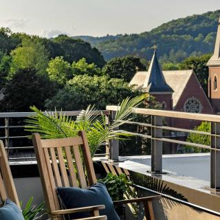 A serene balcony with wooden chairs overlooking a scenic view of trees, hills, and a church in the distance.