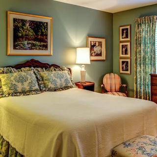 A cozy bedroom with classic decor, featuring a comfortable bed, side table with lamp, paintings on the wall, and floral curtains.