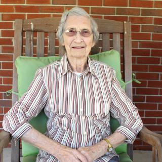 An elderly woman sitting in a wooden rocking chair against a brick wall, wearing a striped shirt and glasses.