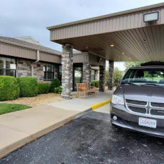 A view of the entrance to a building, featuring parked cars and landscaped surroundings under a cloudy sky.