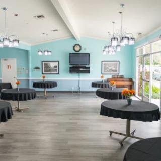 A spacious event room with round tables covered in black tablecloths, decorated with floral centerpieces, and lit by multiple ceiling lights.