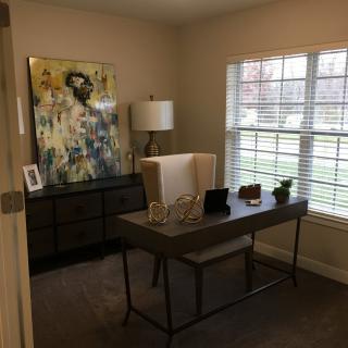A cozy home office with a modern desk, chair, and large window letting in natural light. Artwork and decorations add a touch of elegance.