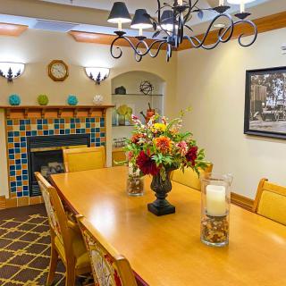 A warmly decorated dining room with a long wooden table, surrounded by chairs and adorned with a floral centerpiece. The room features a colorful tiled fireplace, elegant chandelier, and cozy ambiance.