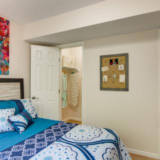 A cozy bedroom featuring blue patterned bedding, vibrant artwork on the wall, and a glimpse into an adjacent closet through an open door.