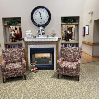 A cozy living room interior featuring a fireplace with two patterned armchairs, a large wall clock above, and decorative items on shelves.