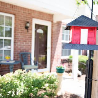 A suburban porch with a red birdhouse and outdoor seating, surrounded by greenery.
