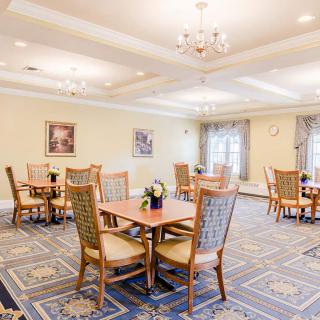 A well-lit dining area with wooden tables and chairs, decorated with chandeliers and patterned carpet.