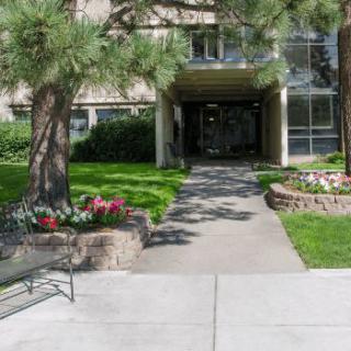 A well-landscaped building entrance with a sidewalk and benches surrounded by flowers and trees.