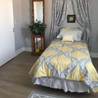 A cozy bedroom with a single bed featuring yellow patterned bedding, a decorative mirror, and a plant on a wooden stand.