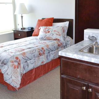 A cozy bedroom with a single bed, featuring floral and orange bedding next to a small sink and lamp.