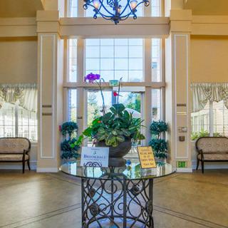 A well-decorated lobby with a central table displaying plants and surrounded by benches, under a large chandelier.