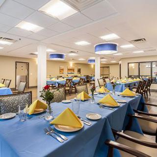 A spacious dining room with tables set for a formal meal, featuring blue tablecloths and yellow napkins.