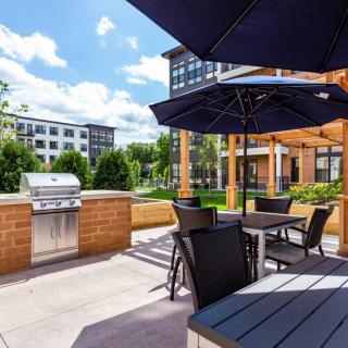 A sunny outdoor patio area with seating, umbrellas, and a BBQ grill in an apartment complex.