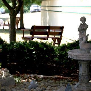 A serene garden scene featuring a bench surrounded by lush greenery and a decorative fountain with a statue.