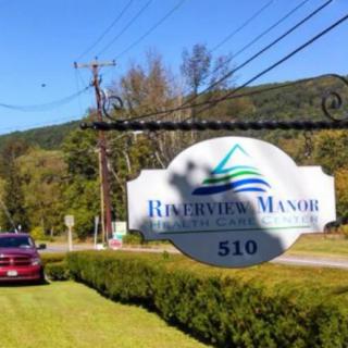 A roadside sign for 'Riverside Manor' surrounded by scenic greenery and mountains.