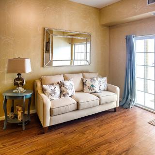 A cozy living room with a beige sofa, decorative pillows, and a side table with a lamp, featuring wooden flooring and a large window.