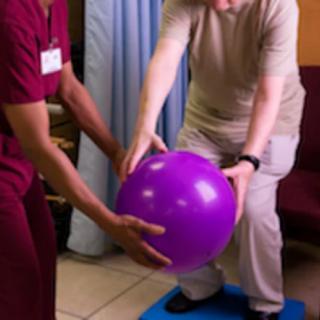 An elderly person is engaged in a physical therapy session, assisted by a healthcare professional, using a purple exercise ball for balance training.