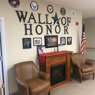 A Wall of Honor display featuring military emblems, photographs, and plaques with an American flag, flanked by two leather chairs and a decorative fireplace.