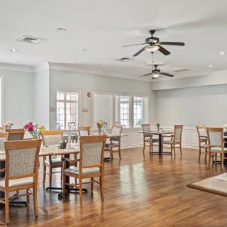 A bright dining room with wooden floors, multiple tables and chairs, ceiling fans, and large windows.