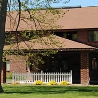 A brick building with a sloped roof, surrounded by grass and trees, featuring a white picket fence.