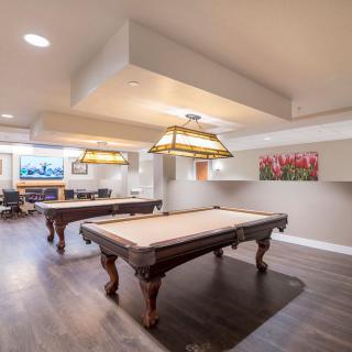 A spacious game room featuring two billiard tables under stylish hanging lights, with wood flooring and a cozy atmosphere.