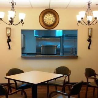 A cozy dining area featuring a central table surrounded by chairs, illuminated by elegant chandeliers and decorated with a wall clock.
