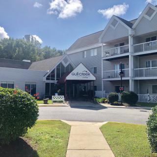 The image shows a residential apartment building with a landscaped entrance on a sunny day.