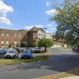 A brick building with a parking lot in front, surrounded by trees under a partly cloudy sky.