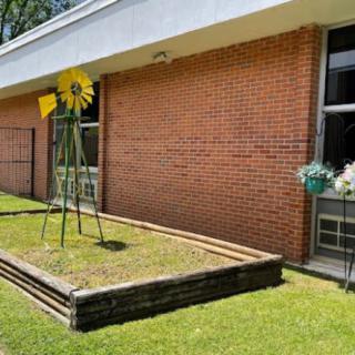 A metal sculpture resembling a sunflower stands in a raised garden bed beside a brick building.