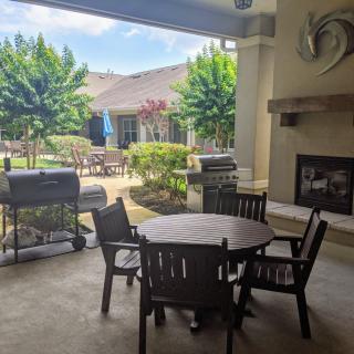 A cozy patio with a circular table and chairs, featuring a fireplace and grill, overlooking a sunny garden.