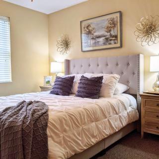 A cozy bedroom with a neatly made bed, featuring tufted headboard and decorative pillows, flanked by nightstands with lamps and wall artwork.