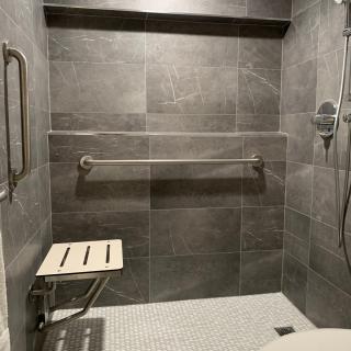 A modern, accessible shower with grey tiles, equipped with a foldable seat and grab bars for safety.