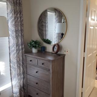 A bedroom corner featuring a wooden dresser with a round mirror, a small plant, and decorative items. Natural light streams in through the window with patterned curtains.