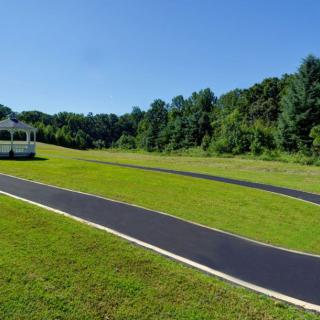 A paved walkway curves around a well-maintained lawn with a gazebo in the background, surrounded by trees under a clear blue sky.