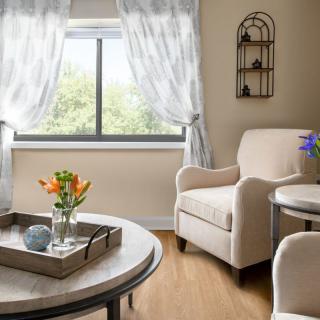 A cozy living room with a comfortable armchair and a coffee table adorned with flowers, set against a sunny window with sheer curtains.
