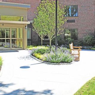 A landscaped entrance featuring a brick building, with a bench and pathway surrounded by trees and shrubs.