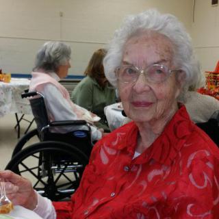 An elderly woman wearing a red outfit is seated indoors, holding a small object.
