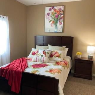 A cozy bedroom featuring a bed with floral bedding, a nightstand with a lamp, and a colorful flower painting on the wall.