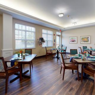 A cozy dining room with neatly arranged tables and chairs, featuring large windows and warm lighting.