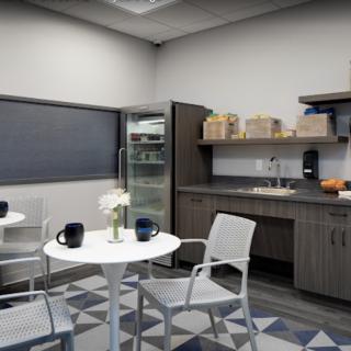 A modern break room with a small kitchenette, featuring tables, chairs, and a refrigerator.