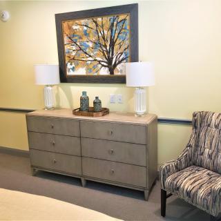 A cozy bedroom setup featuring a dresser with two lamps, an abstract painting, and an upholstered chair.