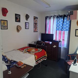 A neatly arranged bedroom with military-themed decor, featuring an American flag curtain, hats on the wall, and a TV on a dresser.